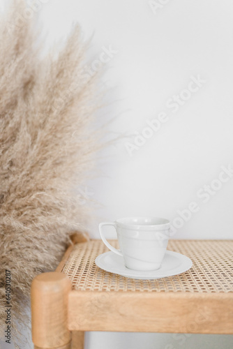A white coffee cup on a saucer on a rattan bench with pampas gra