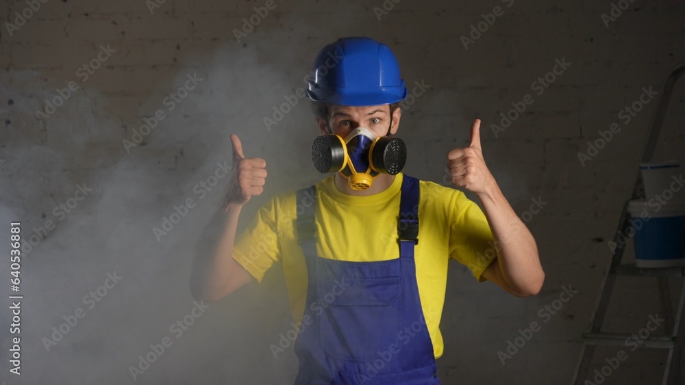 Medium shot of a construction worker standing in the room filled with smoke, happy to wear a respirator and giving a thumbs up.