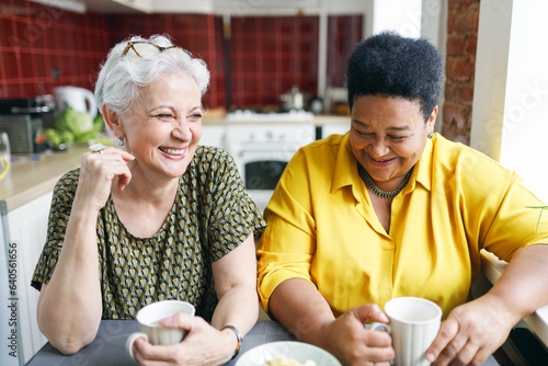 Image of two aged female neighbors of caucasian and african ethnicity drinking coffee together in kitchen having fun sharing gossips and rumors, laughing out loud sitting at table