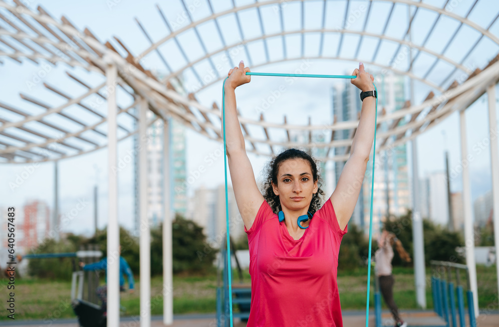 Obraz premium Calm young hispanic woman in sportswear training at sport court using rubber expander looks at camera confidently. Curly caucasian female student at cardio exercise outside. Sport, fitness, health.