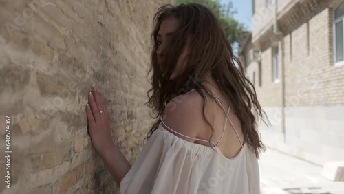 A young woman with flowing long hair and a white sundress walks along an ancient wall in an eastern city photo