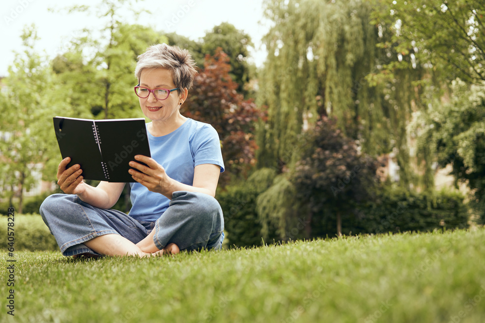 Senior woman in eyeglasses reading book while sitting on grass in park and having a rest Senior woman in eyeglasses reading book while sitting on grass in park and having a rest