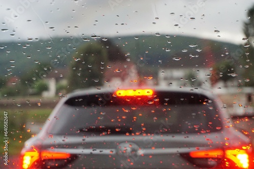Regentropfenmuster auf Glasscheibe vor Auto mit orange-roten Lichtern auf Fähre über Fluss und vor Ortschaft mit Häusern, Wald und Himmel bei Regen am Nachmittag im Herbst