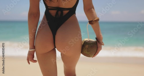 Perfect buttocks of an attractive tanned brunette beach girl holding a coconut drink with a straw. Young woman in a black bikini on the tropical beach. Perfect travel vacation concept.