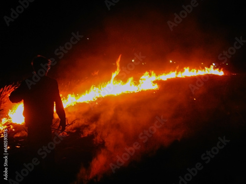 Silhouette of a guy standing against the background of burning grass at night 