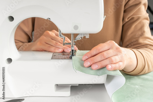 Tailor hands stitching green fabric on modern sewing machine at workplace in atelier. Women's hands sew pieces of fabric on sewing machine closeup. Handmade, hobby, repair, small business concept