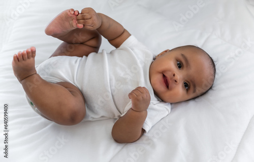 Top view of Happy cute little African newborn baby lying down smiling and looking at camera on white bed in bedroom at home.