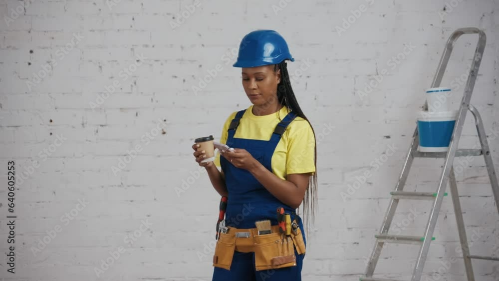 Medium video of a dark-skinned young female construction worker standing in the room under renovation, holding a smartphone and a cup, drinking coffee. Medium video of a dark-skinned young female construction worker standing in the room under renovation, holding a smartphone and a cup, drinking coffee.