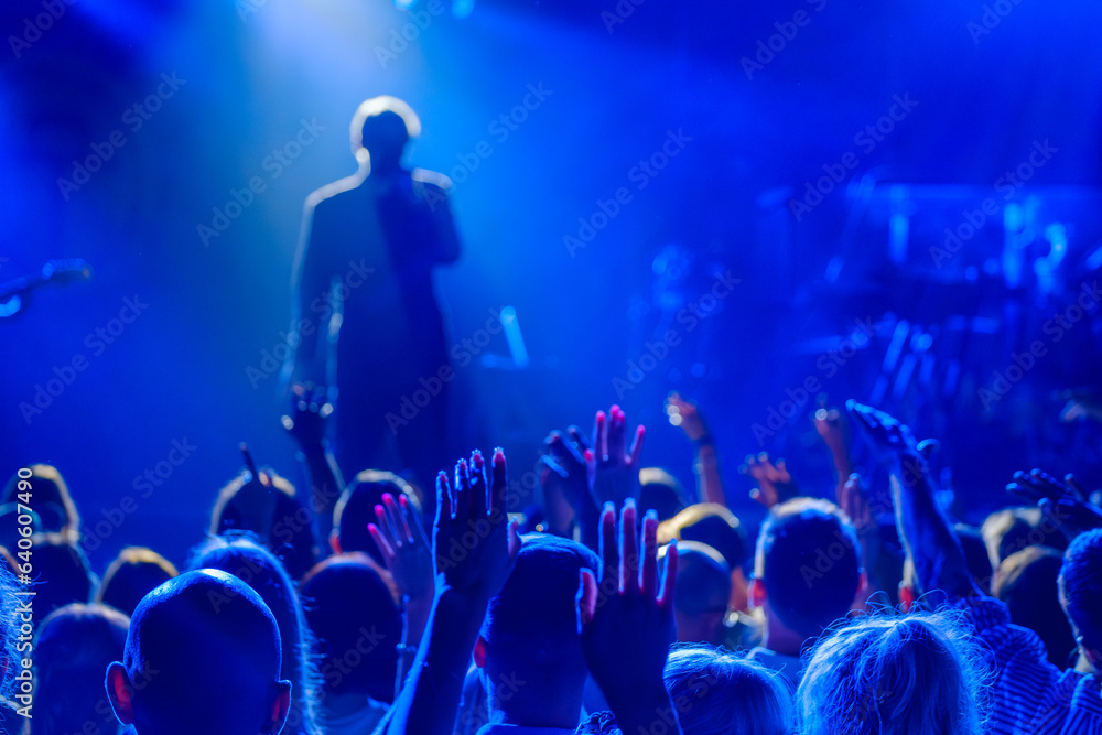 Back view of crowd of unrecognizable people dancing with raised arms while standing near stage during concert of male singer and musician in club
