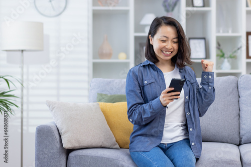Woman celebrating victory at home looking at phone screen, satisfied asian woman happy received online notification of good achievement results sitting on sofa in living room.