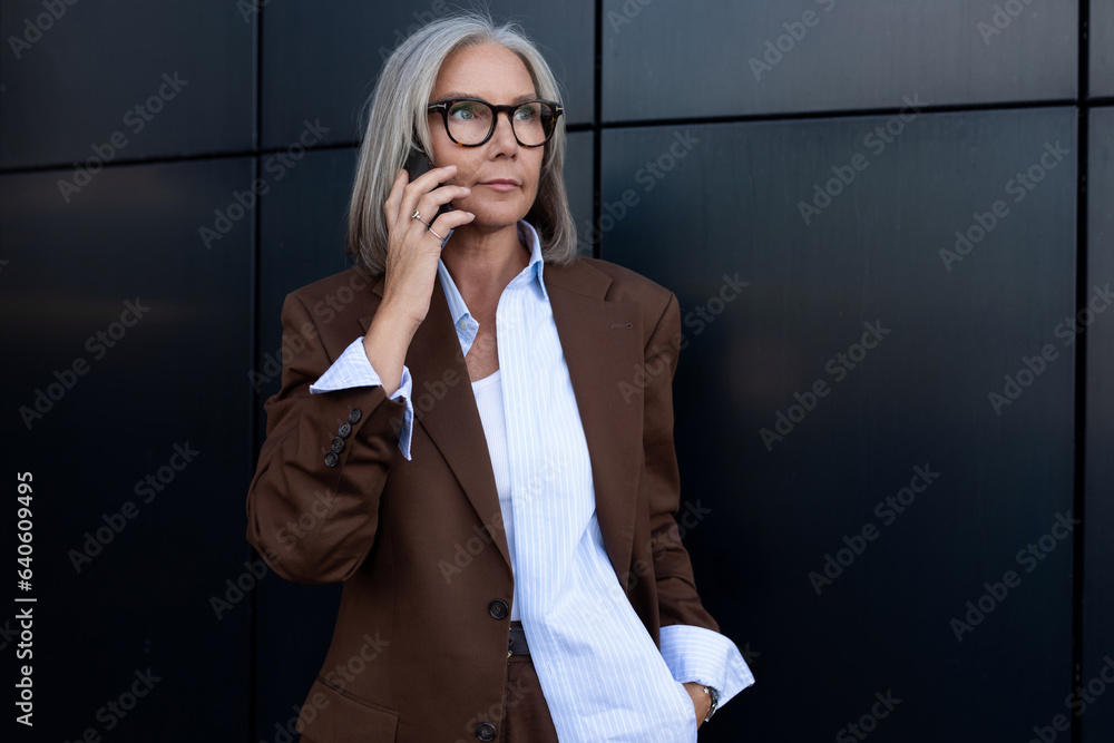 charming elderly gray-haired business woman in a brown jacket and trousers uses a smartphone to discuss business on the street charming elderly gray-haired business woman in a brown jacket and trousers uses a smartphone to discuss business on the street