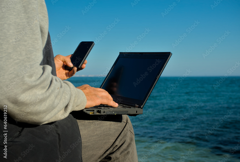 A man with a laptop and phone in his hand sits on the seashore and works. Remote remote work from anywhere in the world A man with a laptop and phone in his hand sits on the seashore and works. Remote remote work from anywhere in the world