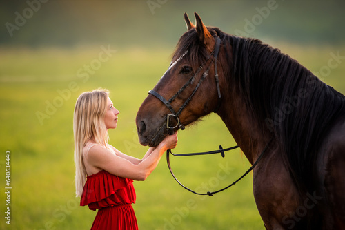 Portrait of a young woman in a red dress walking with a horse in the soft wind light. © Maria Moroz