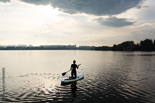 Water sports, surf lessons, the appearance of a girl against the backdrop of a sunrise. Stand on a kayak on the water with the warm colors of a summer sunset. © Artsaba Family