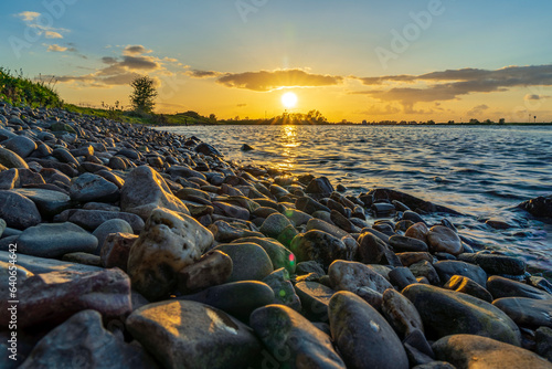 The setting sun shines on the water and on the large pebbles along the bank of the river De Lek near Beusichem, the Netherlands © Emma