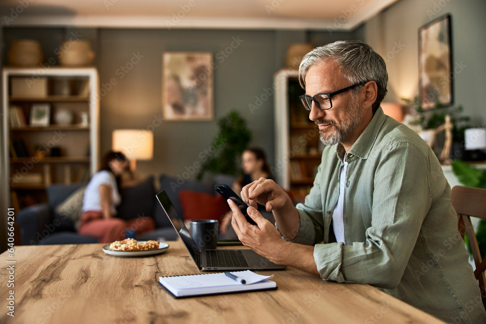 A senior male freelancer typing on a phone while working on a laptop in the living room. A senior male freelancer typing on a phone while working on a laptop in the living room.
