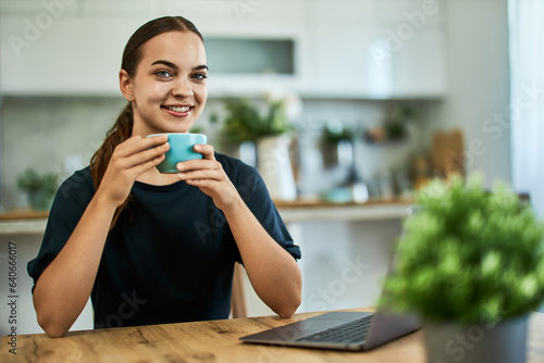 Portrait of a smiling female freelancer holding a cup of tea and working on a laptop.