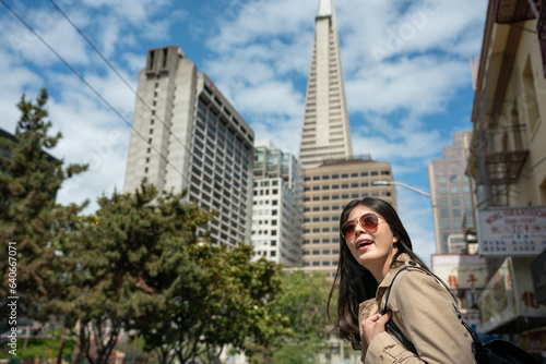 amazed asian Taiwanese woman tourist admiring streetscape with Transamerica Pyramid looming up among tall buildings against blue sky at background in san Francisco's Chinatown
