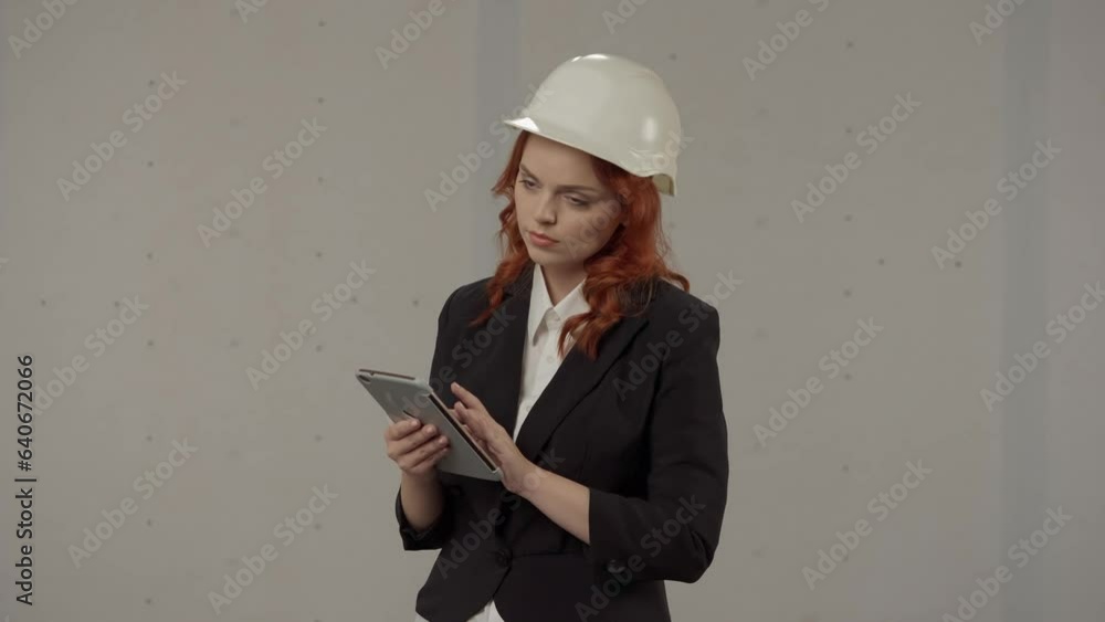 A woman architect conducts an inspection of the object and takes notes in the tablet. Business woman in a protective helmet and with a tablet on a gray background in the studio. A woman architect conducts an inspection of the object and takes notes in the tablet. Business woman in a protective helmet and with a tablet on a gray background in the studio.