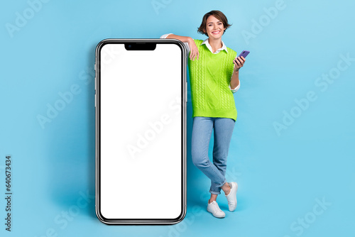 Full size photo of adorable positive girl with brown hairdo wear green vest lean wall hold smart phone isolated on blue color background