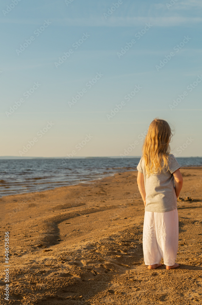 Little cute girl with blond hair standing on the seashore barefoot at sunset and looking into the distance. Local travel trend, authentic tourism