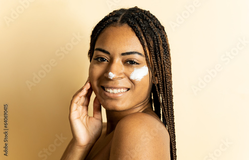A portrait of an African woman with braided hair posing happily on a brown background while looking at the camera. The woman has cream on her nose and left cheek. Relaxing with skin care.