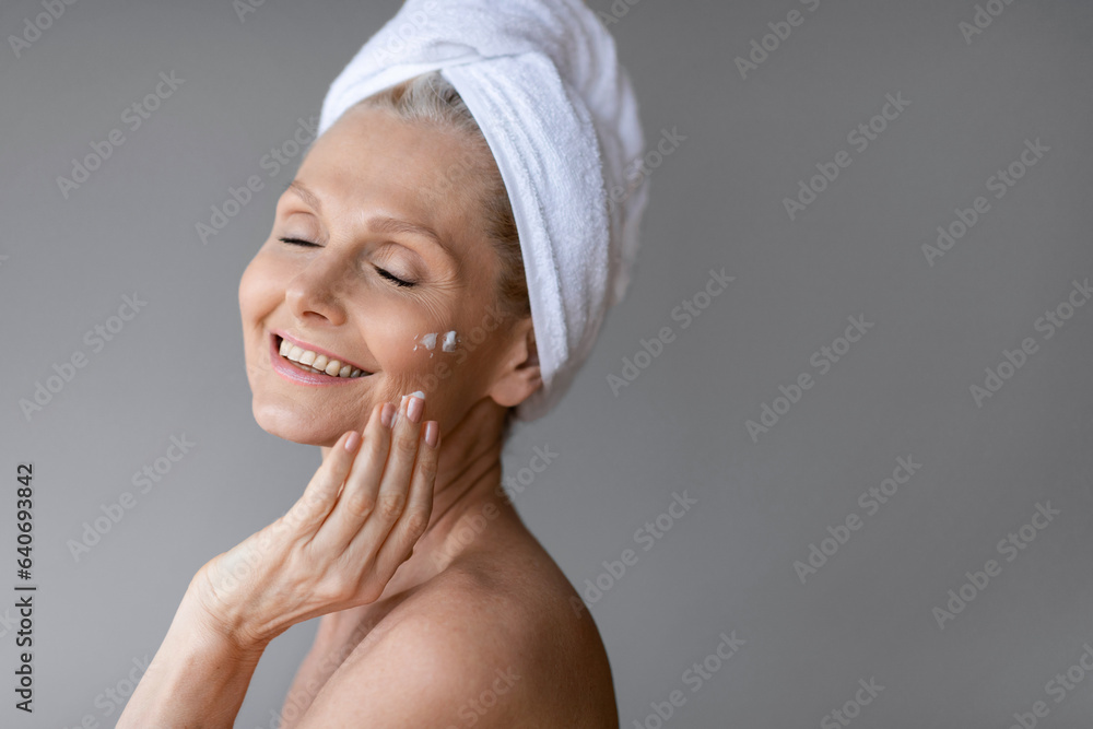 Happy middle aged woman posing with towel on head, applying facial cream on cheek, grey background, copy space Happy middle aged woman posing with towel on head, applying facial cream on cheek, grey background, copy space