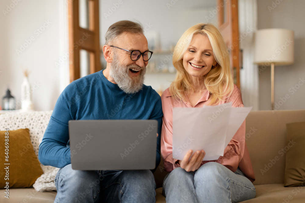 Happy couple of pensioners using computer holding papers at home Happy couple of pensioners using computer holding papers at home