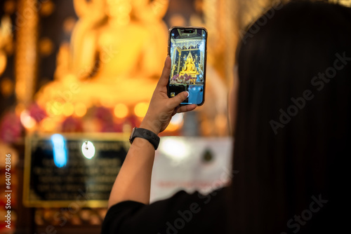 Asian woman tourist is taking picture by cell phone to Buddha for worship with faith to beautiful the golden Buddha images hall in the temple thailand belief in Buddhism.