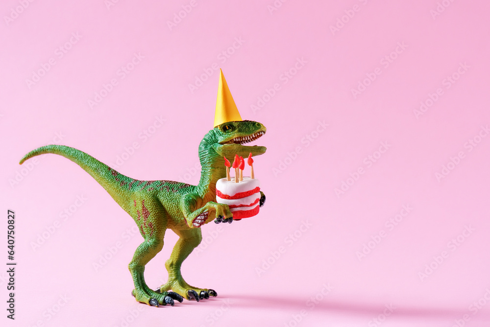 Obraz premium Cute happy green dinosaur in birthday hat holding cake with flaming candles on pastel pink background. Copy space. Minimal art birthday card idea.