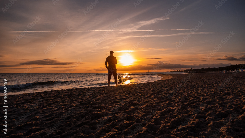 A man standing in front of the sunrise at the Baltic Sea. Next to the man is a camera on a tripod. Sandy beach in Kolobřeh. Kołobrzeg is city in Poland.