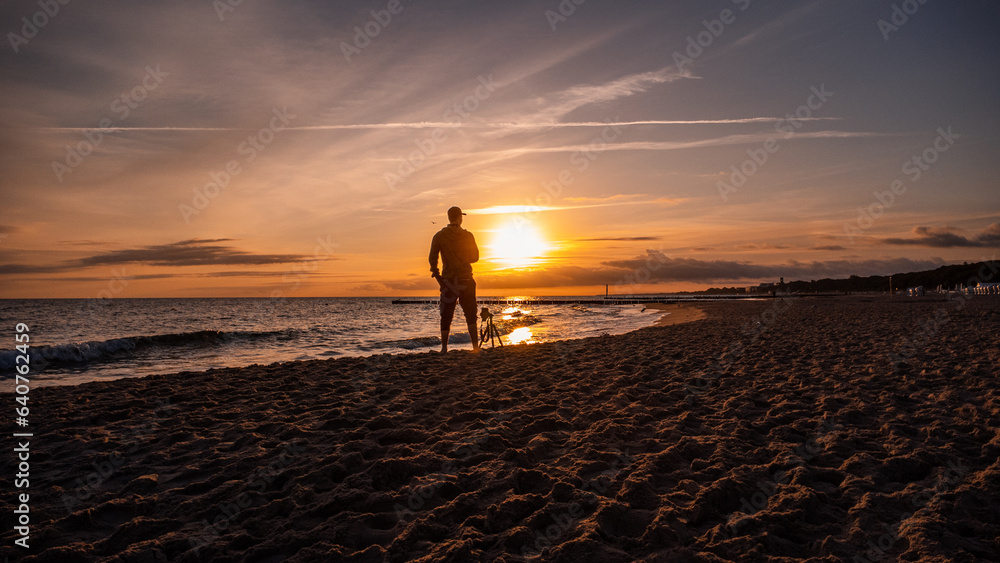 A man standing in front of the sunrise at the Baltic Sea. Next to the man is a camera on a tripod. Sandy beach in Kolobřeh. Kołobrzeg is city in Poland.