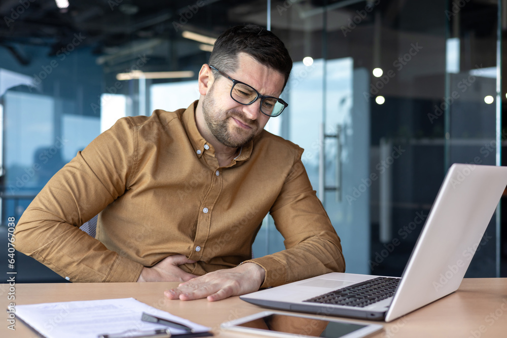 Unwell at work. Young male office worker, businessman sitting in the office and holding his stomach, grimaced in pain and discomfort Unwell at work. Young male office worker, businessman sitting in the office and holding his stomach, grimaced in pain and discomfort