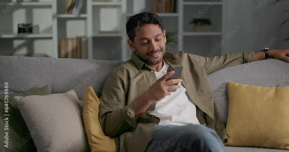 Casually dressed man sitting on sofa, using his smartphone, reading messages. Casually dressed man sitting on sofa, using his smartphone, reading messages.