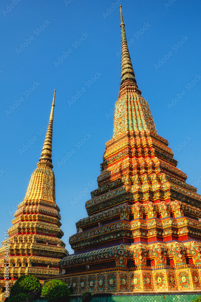 The picture shows ornate temple spires in Thailand, covered in colorful tiles and patterns, lit by the warm glow of the sun.