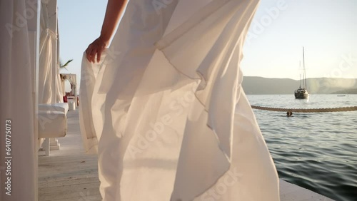 Slow motion of young woman in long flying dress running on wooden sea pier and looking on yachts in susnet light