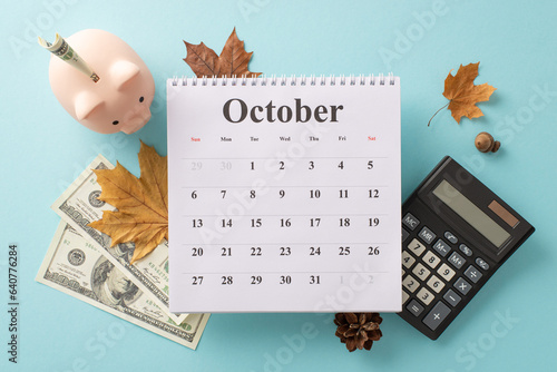 Upswing in expenses and utility payments concept. Top view of piggy bank, dollar bills, calculator, calendar with October month, maple leaves, acorn, pine cone on soft blue surface photo