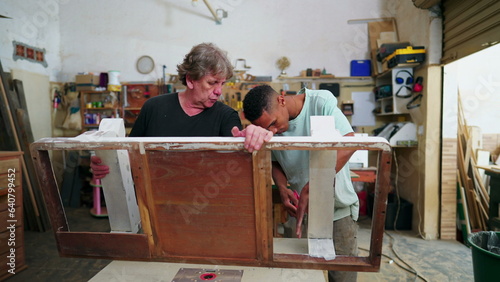 Young and master carpenters engaged with work inside carpentry workshop. Older experienced carpenter guiding employee to polish wood