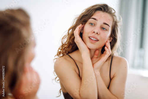 Portrait of a young woman touching a pimple on her face while looking in the mirror. Facial skin problems, medical care and treatment concept.