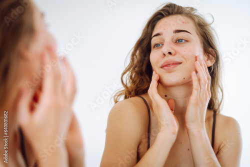 Portrait of a young woman touching a pimple on her face while looking in the mirror. Facial skin problems, medical care and treatment concept.
