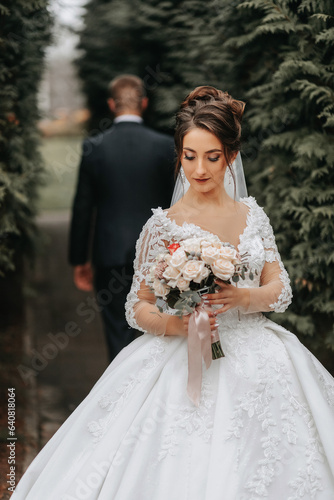Groom and bride walking among tall green trees. Autumn wide-angle photo. Voluminous wedding dress. Classic suit of the groom