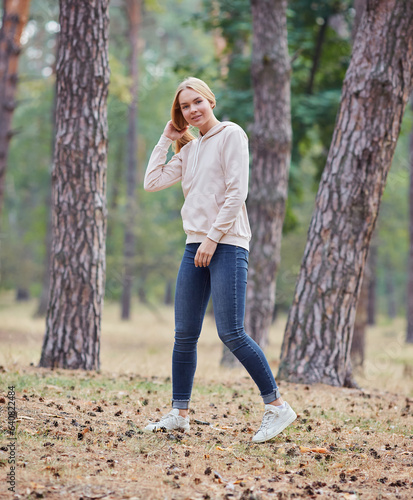 Blue-eyed blonde in a beige hoodie walks in a pine forest. Portrait of a joyful young woman enjoying in autumn park.