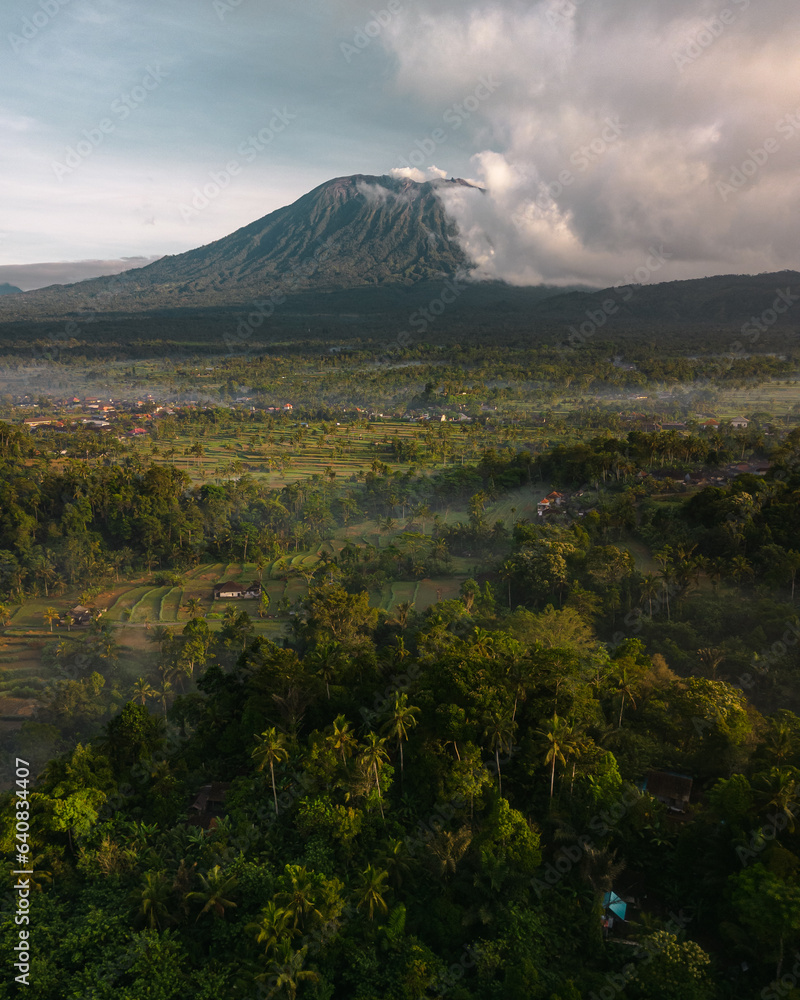 Aerial Vista of Bali's Largest Volcano Mount Agung, Shrouded in Morning Fog, Amidst Palms and Rice Fields. Sunrise in the Countryside. A Vacation Destination in Asia's Indonesia