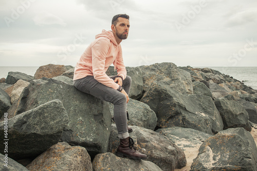 Travel concept. Young bearded man sitting in trendy clothing on the stony shore. Cloudy weather. Text space. Outdoor shot