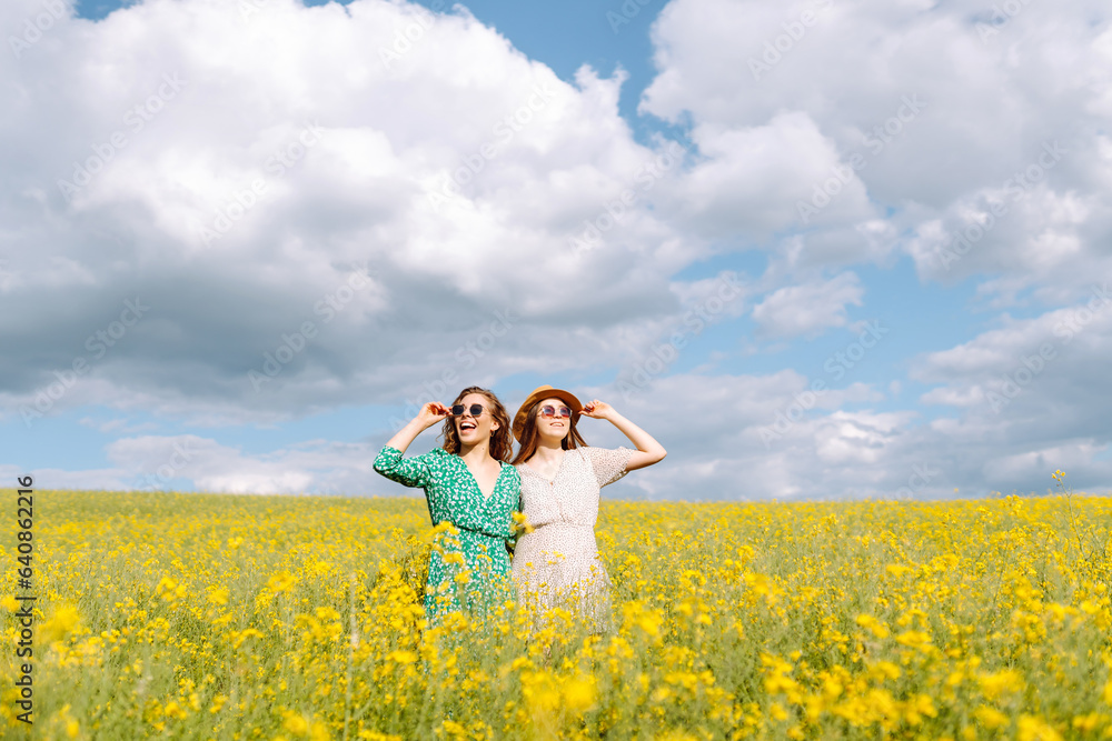 Girlfriends dressed in stylish dresses are walking together in a rapeseed field. Two women enjoy the weather, pose, have fun in a field among blooming rapeseed. Girlfriends dressed in stylish dresses are walking together in a rapeseed field. Two women enjoy the weather, pose, have fun in a field among blooming rapeseed.