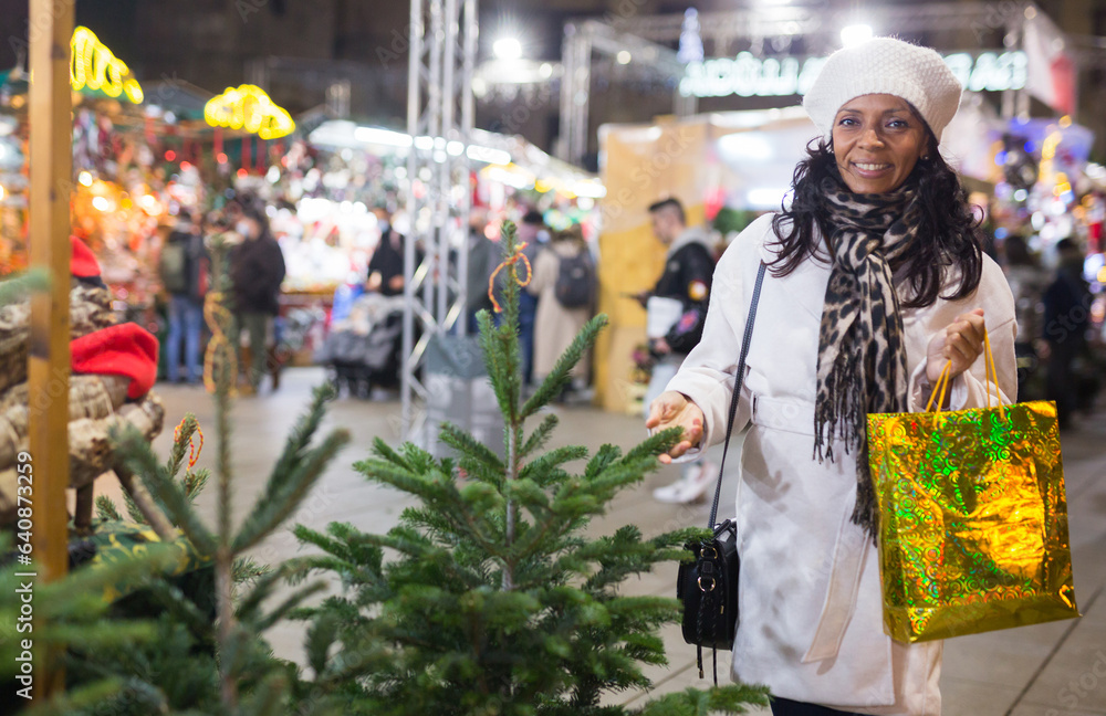 Happy hispanic woman standing near christmas tree at fair and looking in camera.