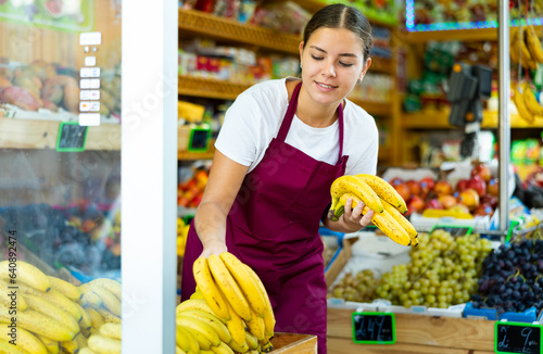 Concentrated young saleswoman working in a vegetable store puts out bananas on the counter for sale
