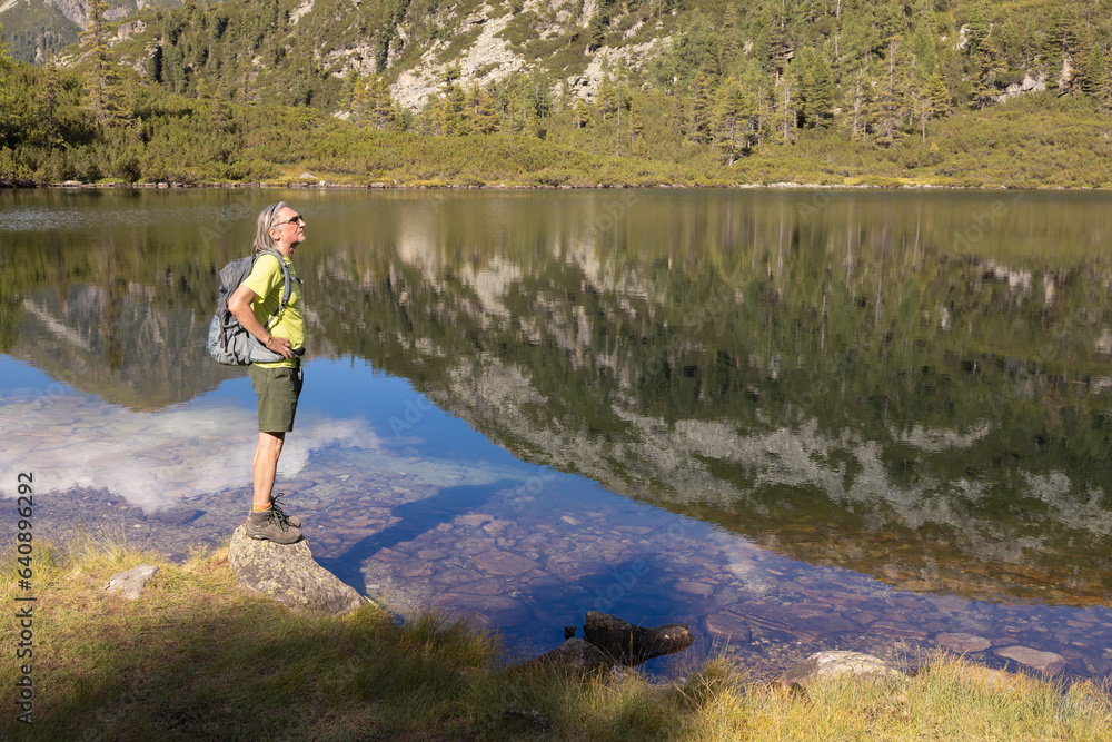 Naklejka premium A man with a backpack stands against the backdrop of a mountain lake with a reflected sky, mountain and clouds