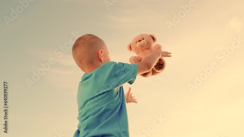 Happy child with teddy bear outdoors on summer day. Little boy throws up and catches bear toy against background of summer nature at sunset. Family weekend in nature. Concept of child's dream to fly