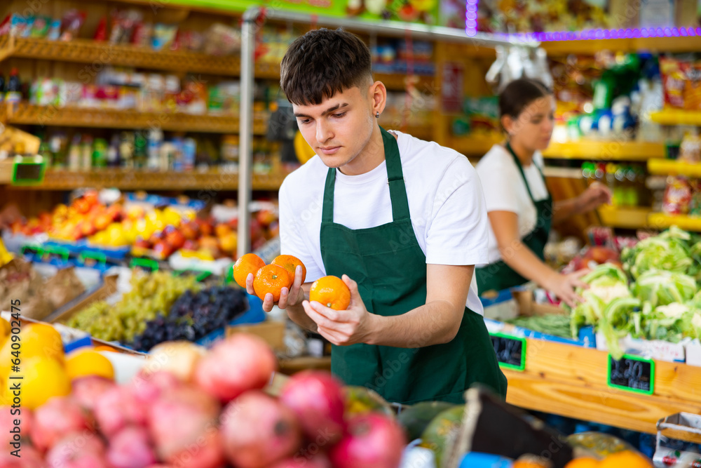 Positive man store employee displaying assortment of oranges at fruit department in supermarket
